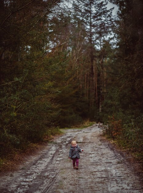 Vertical shot of a little child walking on a muddy road surrounded by trees