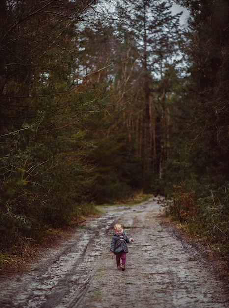 Vertical shot of a little child walking on a muddy road surrounded by trees