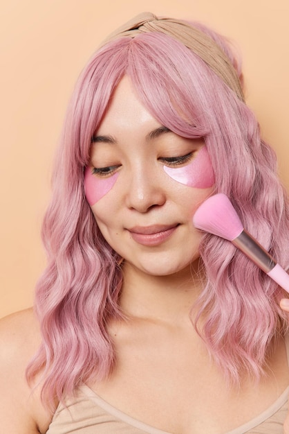 분홍색 머리를 한 사랑스러운 아시아 여성의 세로 샷은 파우더를 바르기 위해 화장용 브러시를 사용하여 베이지색 배경에 대해 붓기를 줄이기 위한 미용 패치를 적용합니다. 일상적인 개념