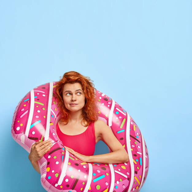 Vertical shot of lovely red haired woman with dimples on cheeks, focused aside, plans future vacation