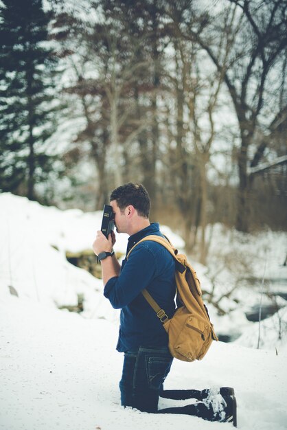 Vertical shot of a male on his knees on snowy ground while holding Bible against his head praying