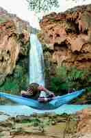 Free photo vertical shot of a male lying on a hammock beside a waterfall flowing down from a hill