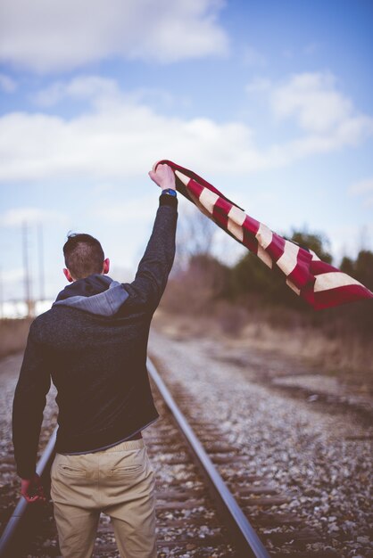 Vertical shot of a male standing on train tracks while holding up the united states flag