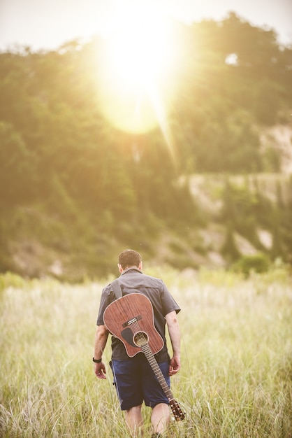 Free photo vertical shot of a male walking in a field next to a forest with a guitar on his back