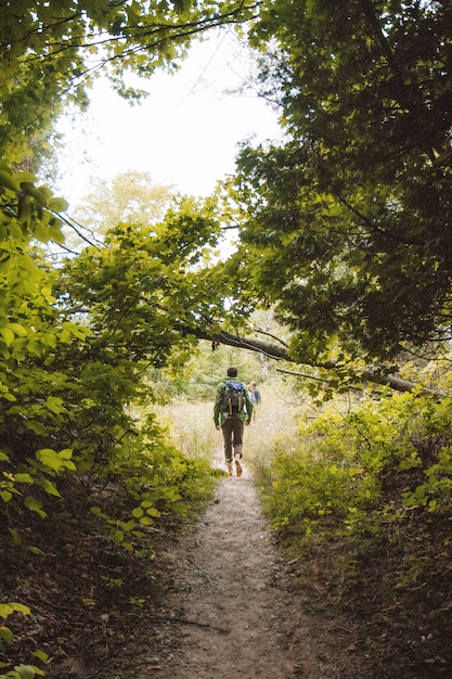 Vertical shot of a male with a backpack walking on a narrow pathway in middle of trees and plants