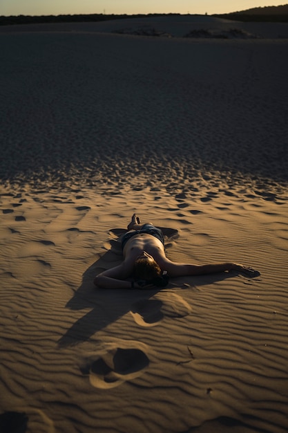 Vertical shot of a man laying on the sand in a deserted landscape at sunset