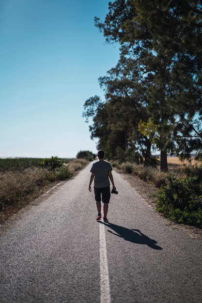 Вертикальный снимок человека с фотоаппаратом, идущего посреди пустой дороги с деревьями сбоку