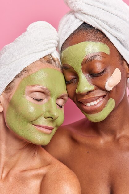 Вертикальный снимок многонациональных женщин разного возраста, которые наносят на лицо зеленые натуральные отшелушивающие маски, проходят косметические процедуры после принятия душа поза с голыми плечами в помещении носят банные полотенца на голове