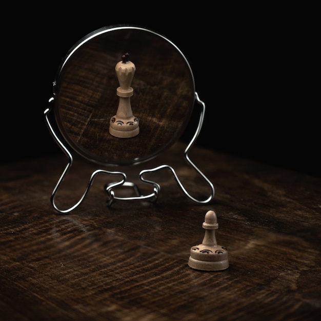 Вертикальный снимок шахматной пешки на деревянном столе и зеркала, отражающего ее как шахматную королеву