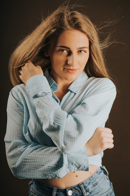 Бесплатное фото Вертикальный снимок привлекательной блондинки в джинсах и короткой рубашке, позирующей на коричневом фоне