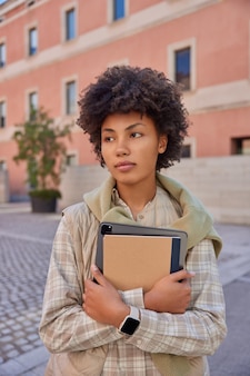 Вертикальный снимок красивой кудрявой женщины с блокнотом и планшетом с приложением для студентов, в клетчатой рубашке и жилете ходит по улицам