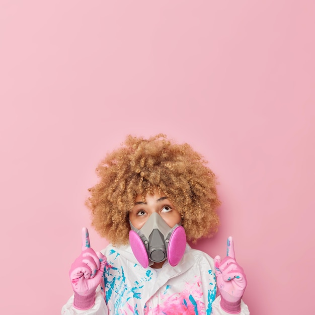 Бесплатное фото Вертикальный снимок кудрявой женщины в противогазе и медицинском костюме биологической опасности указывает вверху на пустое место, показывает место для вашей рекламы, рассказывает о химической защите, изолированной на розовой стене