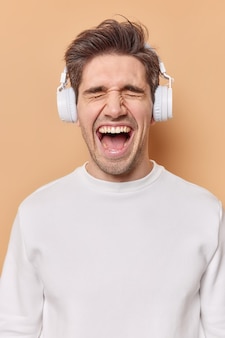 Вертикальный снимок эмоционального молодого человека, который чувствует себя вне себя от радости, держит широко открытым ртом, слушает музыку с громким звуком, носит стереонаушники в ушах, носит повседневный джемпер, изолированный на бежевом фоне.