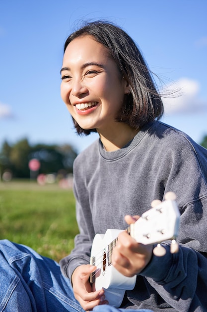 無料写真 公園に座ってウクレレの歌とリラックスの仕方を学ぶ幸せな韓国の女の子の垂直ショット