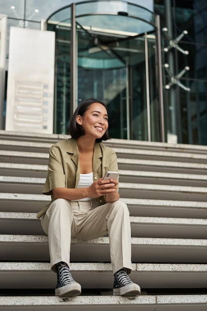 Бесплатное фото Вертикальный снимок счастливой улыбающейся азиатской девушки, сидящей на лестнице в центре города, держит мобильный телефон и смотрит