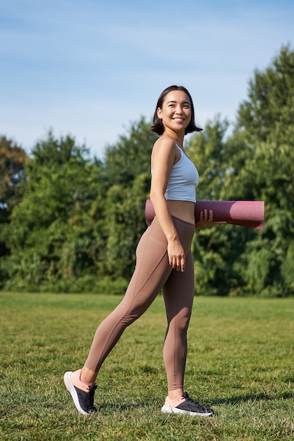 Бесплатное фото Вертикальный снимок стильной фитнес-женщины с резиновым ковриком, позирующей на лужайке в парке, тренирующейся на свежем воздухе