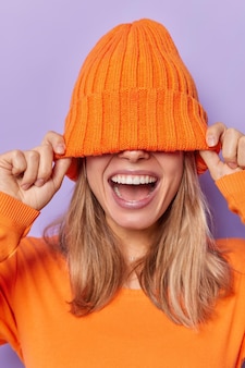 Вертикальный снимок до неузнаваемости женщины, скрывающей лицо в теплой оранжевой шляпе, радостно смеется, держит рот открытым, чувствует себя счастливыми, позирует на фоне фиолетовой стены студии, пытается кого-то развлечь. концепция эмоций