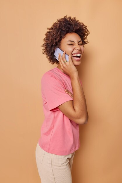 Vertical shot of overjoyed curly haired woman laughs while has telephone conversation discusses something very funny dressed in casual t shirt and trousers stands sideways against brown background