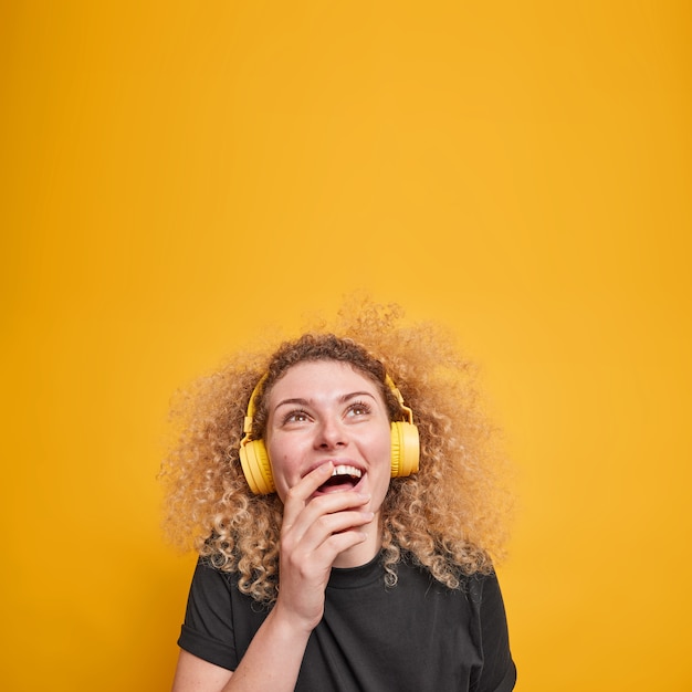 Vertical shot of overjoyed curly haired woman looks above gladfully keeps hand on mouth wears stereo headphones enjoys favorite playlist dressed in black casual t shirt isolated over yellow wall