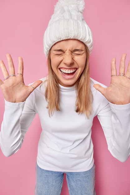 Free photo vertical shot of overjoyed teenage girl wears warm knitted hat jumper and jeans keeps palms raised towards camera has playful mood smiles broadly keeps eyes closed poses against pink background