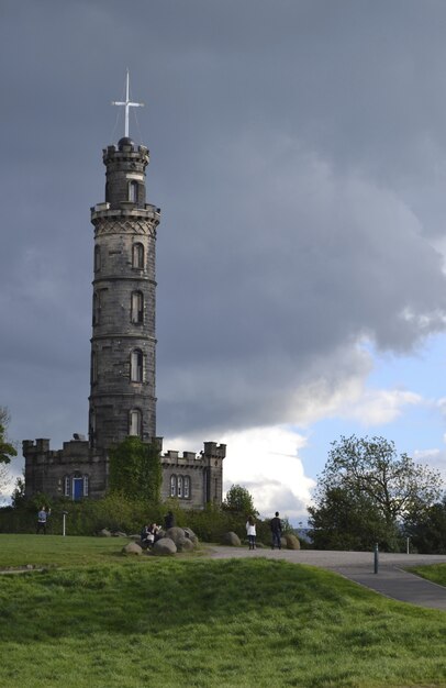 Vertical shot of a pathway with people walking near a grassy field and a tower under a cloudy sky