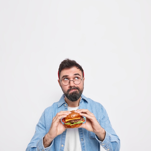 잠겨있는 수염 난 남자의 세로 샷 위에 초점을 맞춘 식욕을 돋우는 햄버거가 뭔가에 대해 깊이 생각합니다.