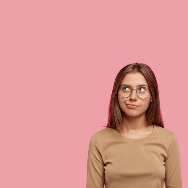 Vertical shot of pensive beautiful young woman concentrated upwards, looks doubtfully and thoughtfully, dressed in casual clothes, has round spectacles, isolated over pink wall.