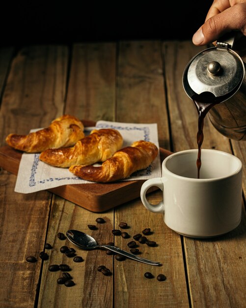 Ripresa verticale di una persona che versa il caffè in una tazza bianca con tre croissant su una tavola di legno