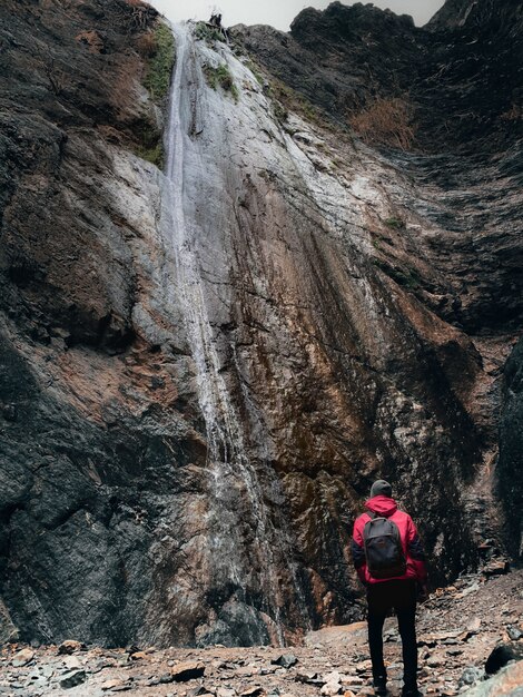 Vertical shot of a person in a red coat and a backpack looking at a high cliff with waterfall