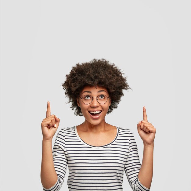 Vertical shot of pleasant looking African American female with joyful expression, has curly hair, points upwards with index fingers