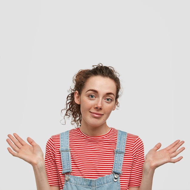 Vertical shot of pleasant looking clueless female with curly hair, has hesitant expression, wears striped t-shirt with dungarees, doesn`t know answer on question, isolated over white wall