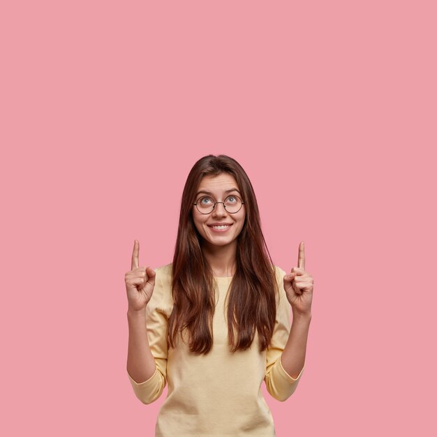 Vertical shot of pleasant looking European woman with positive expression, points upwards with both index fingers, counts stars, likes what she sees