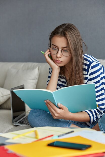 Vertical shot of pleasant looking European young businesswoman looks over some papaerwork, works at home on sofa.