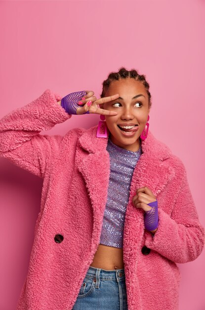 Vertical shot of pleasant looking young woman makes peace gesture, sticks out tongue and looks aside, brings positive vibes, enjoys excellent day off, dressed in pink coat, has cornrow hairstyle