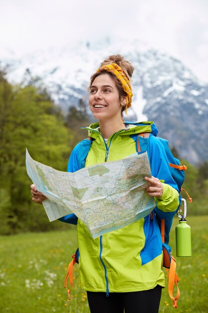 Вертикальный снимок довольной кавказской путешественницы с картой путешествия, одетой в яркий анорак