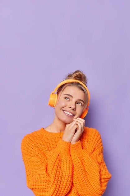 Vertical shot of pleased dreamy woman smiles gently keeps hands near face has pensive expression wears casual knitted sweater listens music via wireless headphones isolated over purple wall