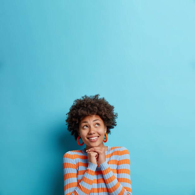 Vertical shot of pleased young Afro American woman keeps hands under chin focused upwards happily dreams about something wears casual striped jumper isolated over blue wall