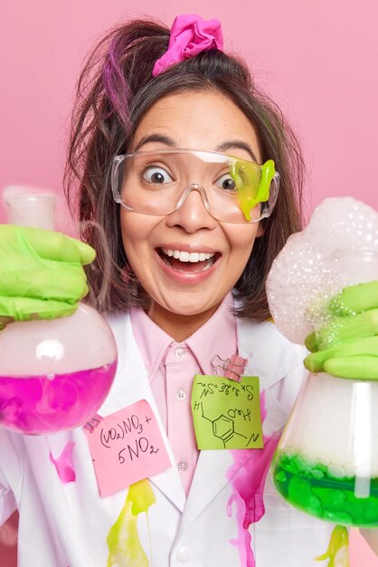 긍정적인 아시아 여성의 세로 샷은 실험실에서 밝은 버블링 액체가 있는 유리 비커 두 개를 들고 기뻐 놀란 표정을 짓고 있습니다. 여성 약사가 신약을 만든다