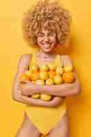 Free photo vertical shot of positive curly haired woman embraces big heap of juicy citrus tropical fruits holds oranges and lemons dressed in swimsuit smiles positively isolated over vivid yellow background