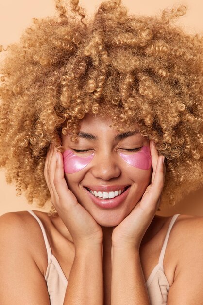 Vertical shot of positive curly haired woman touches face gently applies pink hydrogel patches under eyes to remove wrinkles smiles gladfully keeps eyes closed stands bare shoulders Beauty concept