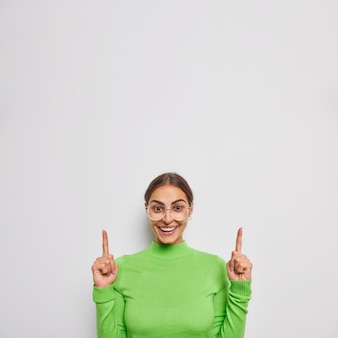 Vertical shot of positive excited young woman points fingers up points above at logo or store advertisement wears eyeglasses and green turtleneck isolated over white wall
