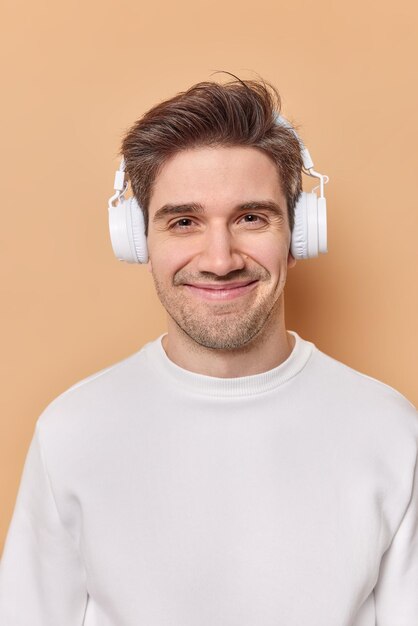 Vertical shot of positive male teenager smilespleasantly has good mood listens music via headphones feels pleased uses stereo headphones wears casual white jumper isolated over brown studio wall