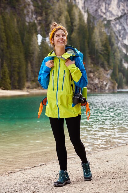 Vertical shot of positive traveler strolls across shore near mountain lake, carries camera