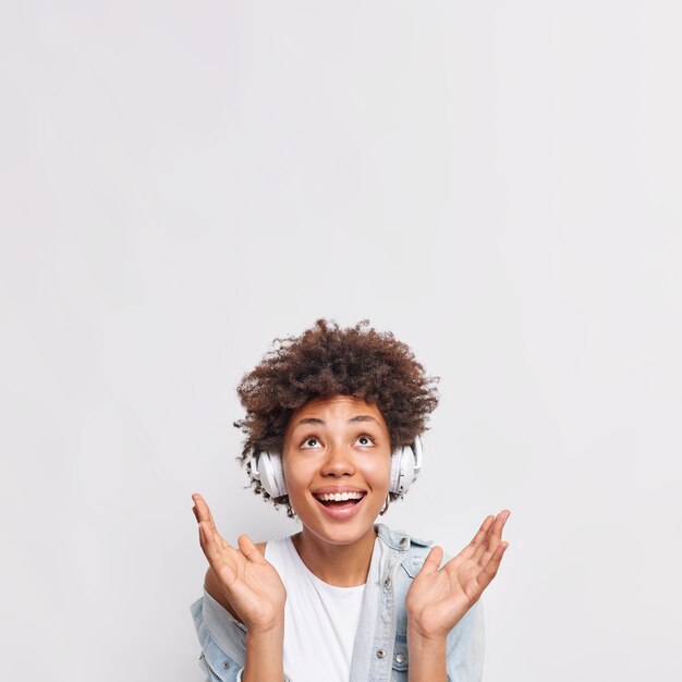 Vertical shot of positive woman spreads palm focused above enjoys perfects sound in wireless headphones has fun listens music for free isolated over white wall