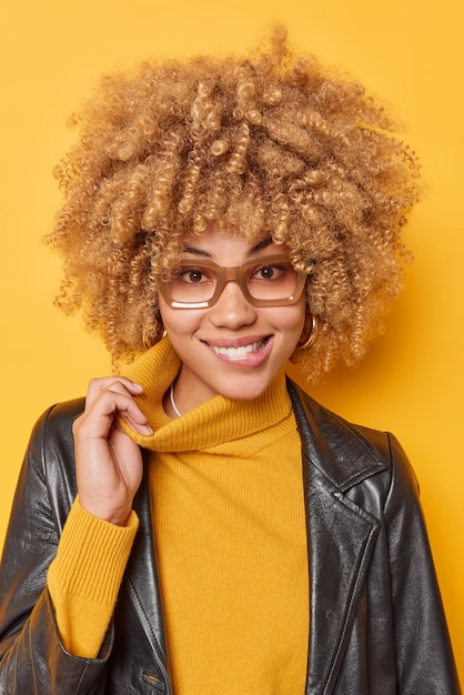 ポジティブな若い女性モデルの垂直ショットは、タートルネックの唇を伸ばし、眼鏡をかけています。革のジャケットは、黄色の背景の上に孤立して幸せを感じます。ファッショナブルな縮れ毛の女性