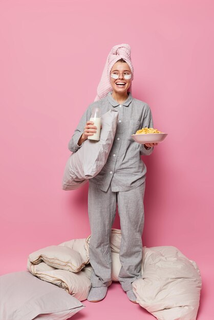 Vertical shot of positive young woman dressed in pajama towel on head has healthy breakfast holds bowl with cornflakes and milk undergoes beauty procedures after sleeping enjoys great morning at home