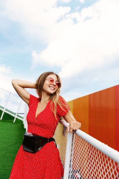 Vertical shot of a pretty blond girl in summer dress and red sunglasses, leaning on handrail in park.