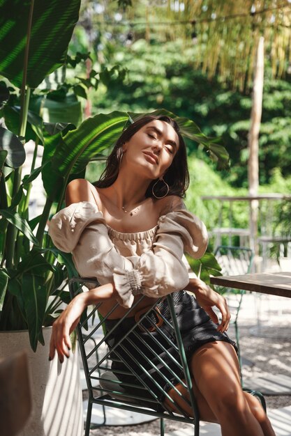 Vertical shot of pretty brunette woman sitting in shadow of tropical palm leaves.