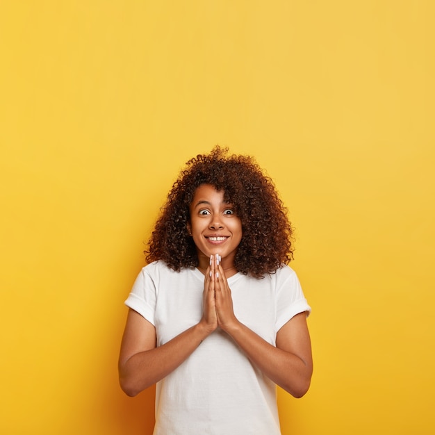 Free photo vertical shot of pretty curly haired teenage girl has surprised happy look, smiles broadly, keeps palms together in praying gesture, believes in gods help, has hopeful expression, wears white clothing