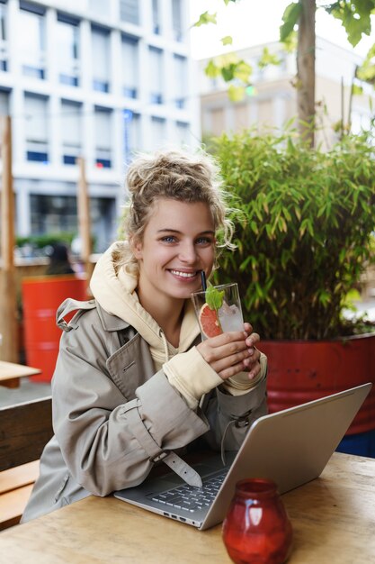Вертикальный снимок довольно улыбающейся женщины, выглядящей счастливой, попивая коктейль, сидя в кафе на открытом воздухе, используя ноутбук.
